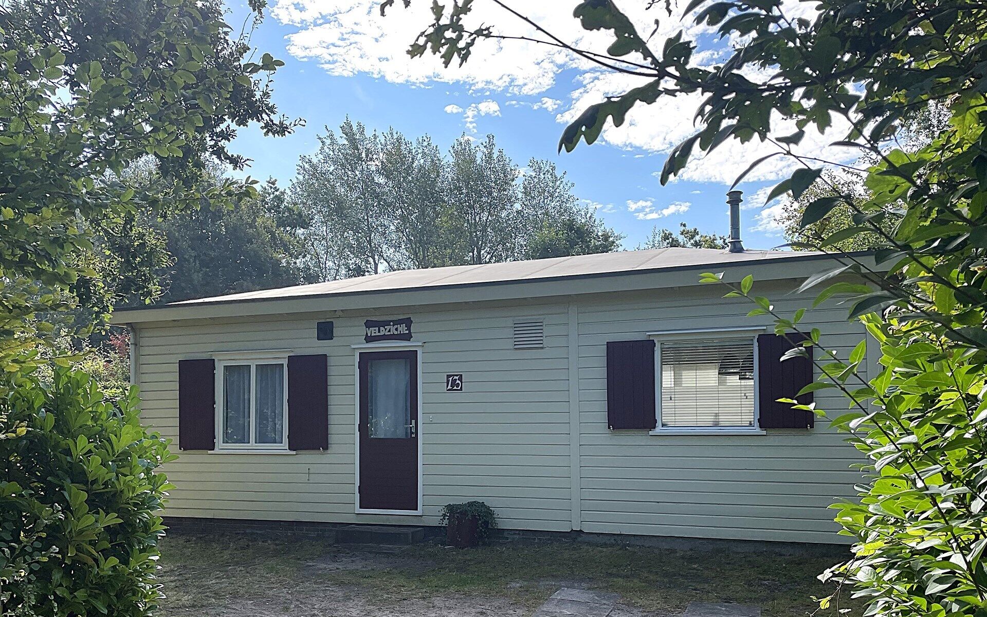 A small, single-story mobile home with a dark brown door and windows, labeled "VELDZICHL" and numbered "13".