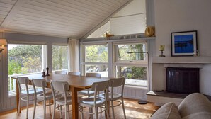 Un coin repas avec une table en bois et des chaises blanches, de grandes fenêtres avec des rideaux, une cheminée et un tableau encadré au mur.