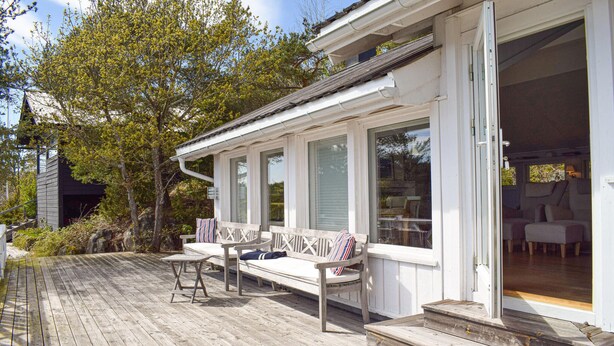Une terrasse en bois dotée d’un espace salon couvert, d’un coin repas visible à travers la porte ouverte et d’une vue sur les arbres environnants.