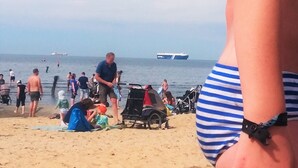 Escena de playa con gente, un cochecito de bebé y un barco al fondo.