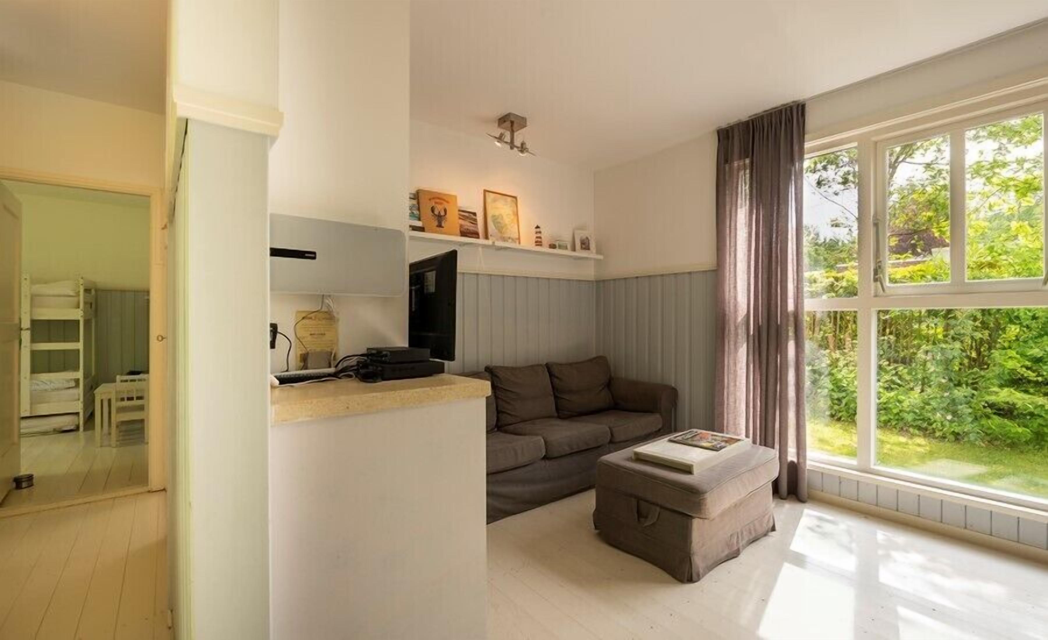 A living room with a sofa, ottoman, and a television on a stand. There is a window with curtains and a view of greenery outside.