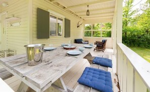 Un espacio exterior techado con comedor de madera para cuatro personas, una olla grande y asientos con cojines azules.