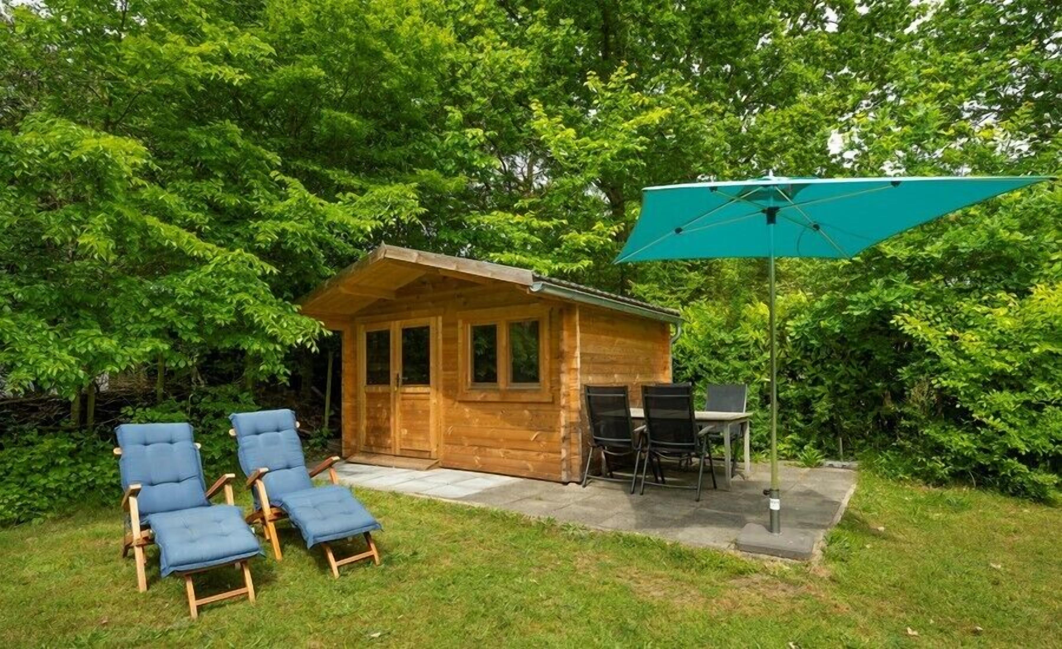 En træhytte med en terrasse, to blå liggestole, et bord og en grøn parasol.