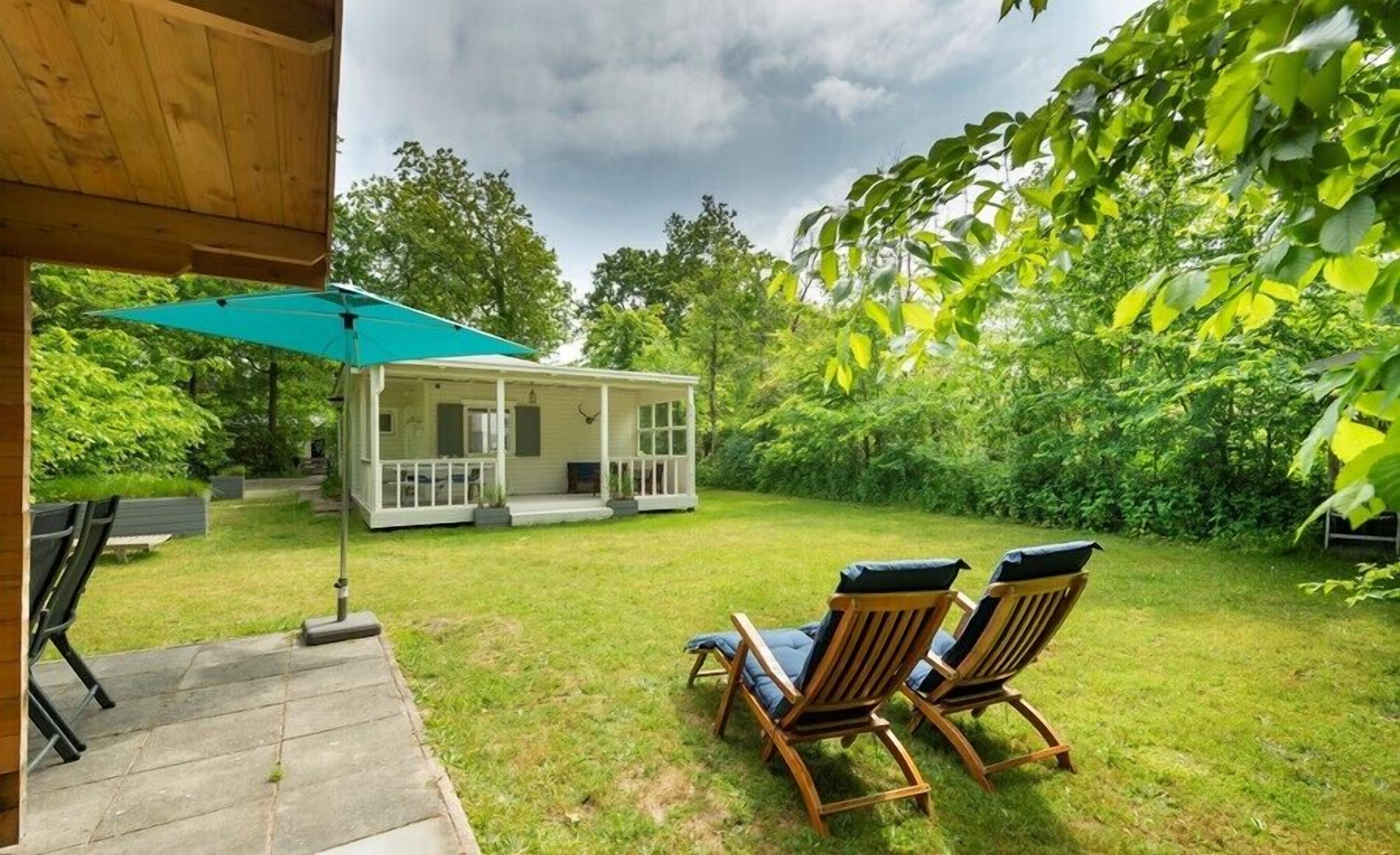 Un patio cubierto con dos sillas de madera y una sombrilla verde, una pequeña casa blanca con porche, y exuberante follaje al fondo.