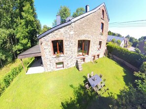Ein Steinhaus mit geneigtem Dach, einer kleinen Veranda und einem grasbewachsenen Garten mit Tisch und Stühlen.