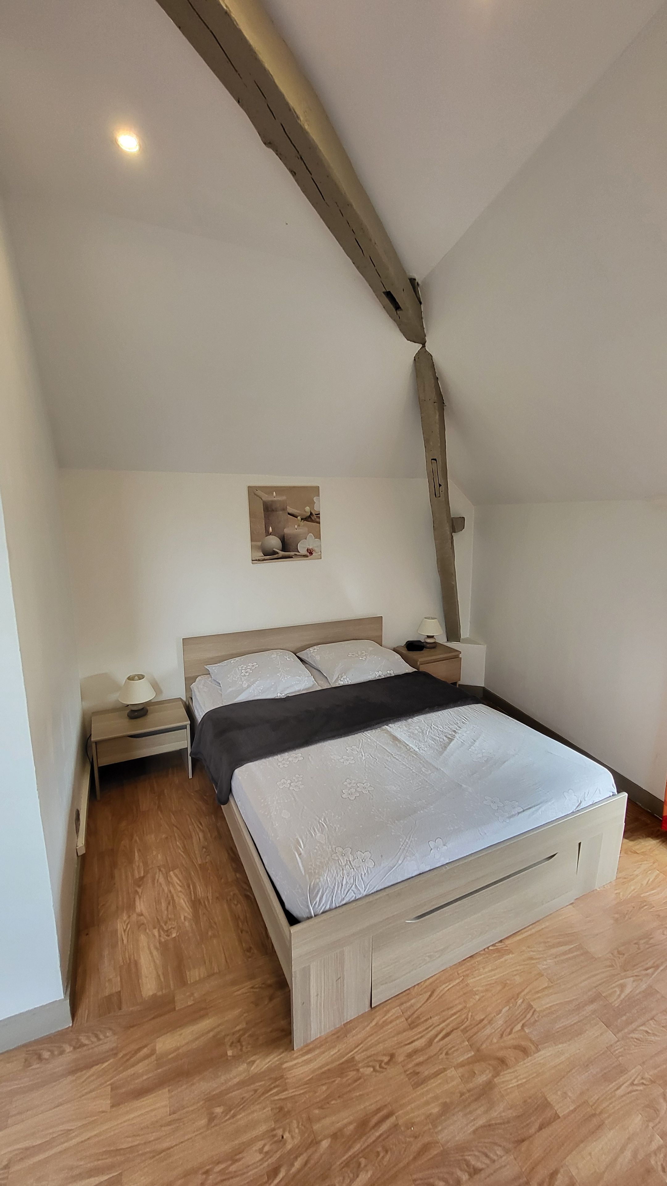 Une chambre à coucher avec un lit en bois, une table de chevet, une lampe fixée au mur et un plafond avec des poutres apparentes.