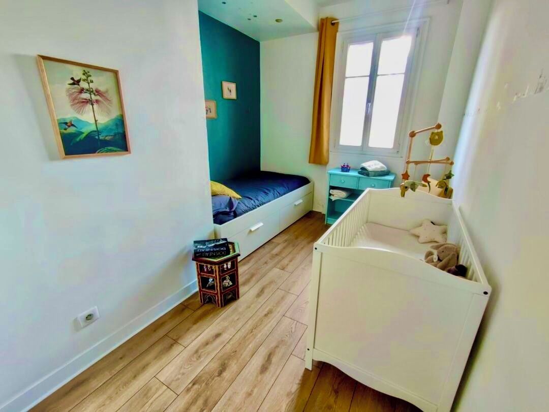 A room with a bed, a crib, a small table, and a window with curtains.