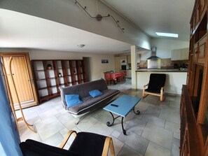 A living room with a sofa, two armchairs, a coffee table, and a dining area with a table and chairs. There is a kitchen visible in the background.