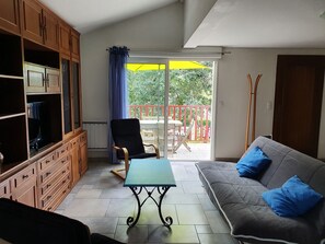A living room with a sofa, a table, and a TV on a cabinet. There is a balcony with a red railing and a yellow umbrella.