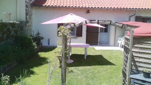 Una casa con techo de tejas rojas, un pequeño jardín con mesa y sombrilla, y una valla de madera.