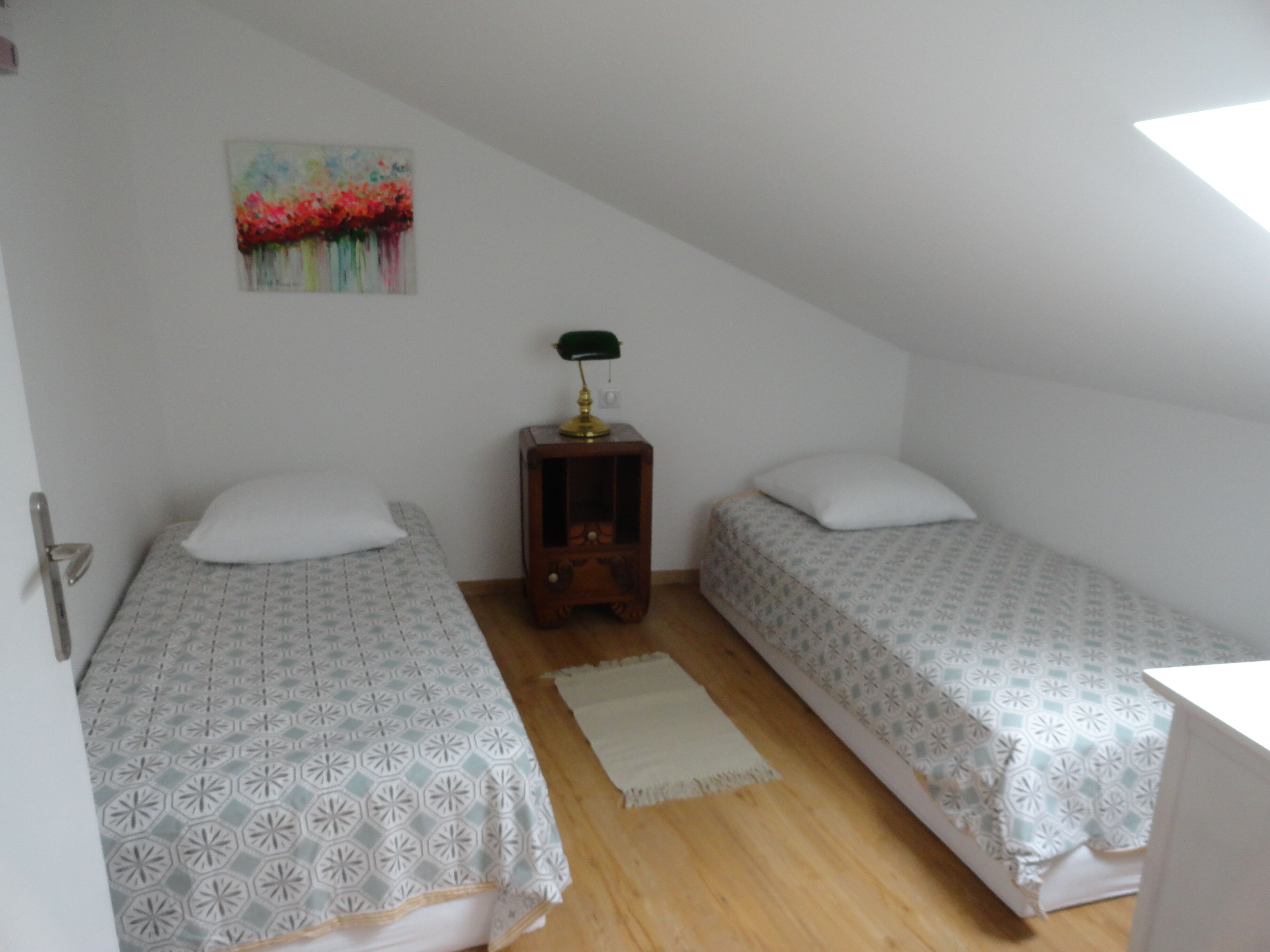 A small, single-room accommodation with two beds, a nightstand, a painting, and a window.