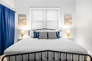 Une chambre à coucher avec un lit, deux tables de chevet avec des lampes, une fenêtre avec des stores et un rideau bleu.