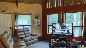 A spacious living room with a leather sofa, a wooden dining table, and a large window with a view of the outdoors. - Cabin in the pines (Munds Park)