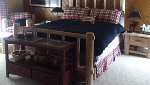 A cozy room with a wooden ceiling, a bed with plaid bedding, a television on the wall, and a small table with a bowl and candles. - Cabin in the pines (Munds Park)