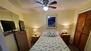A bedroom with a bed, two bedside tables with lamps, a ceiling fan, a TV on a dresser, and a framed picture on the wall. - Newly renovated with the lowest rates you can find. (Hilton Head Island)