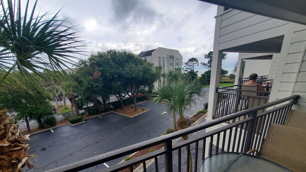 A balcony with a glass table and chair, overlooking a parking lot with trees and a building in the background. - Newly renovated with the lowest rates you can find. (Hilton Head Island)