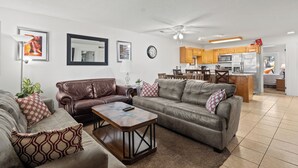 Una sala moderna con un sofá de cuero marrón, un sofá gris y una mesa de centro de madera. Se observa una cocina al fondo con armarios de madera y un microondas.