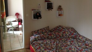 Une chambre avec une couvre-lit à motifs floraux, une petite étagère sur laquelle est posé un ours en peluche, et un miroir qui reflète une chaise et une lampe.