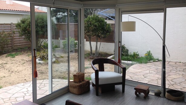 Une pièce dotée d’une porte-fenêtre coulissante qui donne sur un patio. À l’intérieur, on trouve une chaise en bois avec un coussin, une petite table en bois et un panier pour animaux de compagnie.