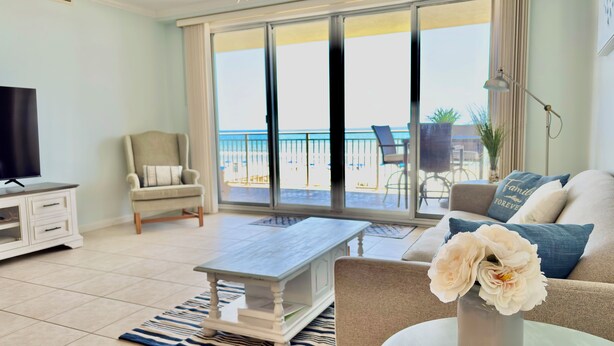 A living room with a TV, a sofa, a chair, a coffee table, and a view of the beach through large glass doors.