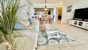A living room with a sofa, coffee table, and a TV. There is a kitchen in the background.