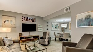 Un salon moderne avec un canapé, un fauteuil et une table basse en verre. Un téléviseur à écran plat est fixé au mur, et on y voit des œuvres d’art encadrées ainsi qu’une enseigne portant l’inscription « SAVANNAH » au-dessus.