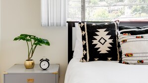 Una camera da letto con un letto, un comodino con una pianta e una sveglia, una finestra con tende e vista sul verde all'esterno.