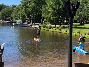 Ein ruhiger Fluss mit Stand-Up-Paddleboards, Booten und einem Park mit Bäumen und Bänken.