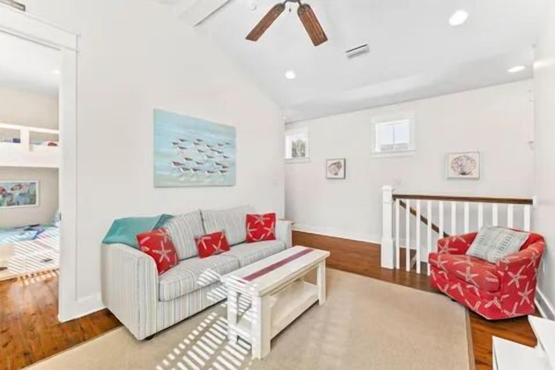 Amplia sala de estar con un sofá, una mesita de centro y un sillón rojo. Se observa una escalera con barandilla y una puerta que da a otra habitación.