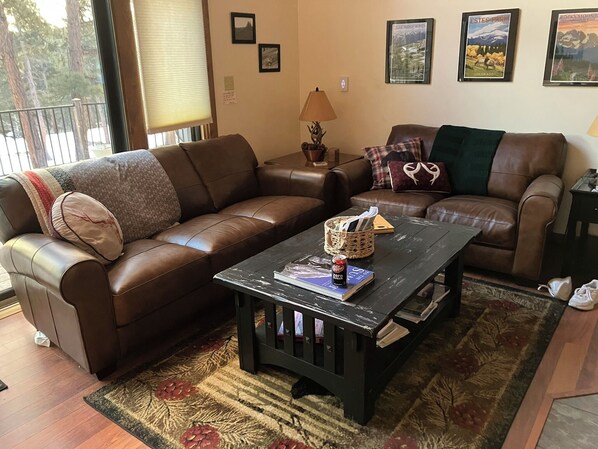 A living room with two brown leather sofas, a wooden coffee table with books and a can, a patterned rug, and framed pictures on the wall.