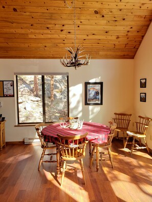 A dining area with a wooden ceiling, a chandelier, and a large window with a view of trees.