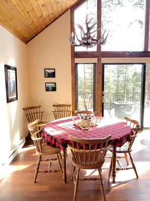 A dining area with a large table covered in a checkered tablecloth, surrounded by wooden chairs. There is a chandelier with antlers above the table, and framed pictures on the wall.