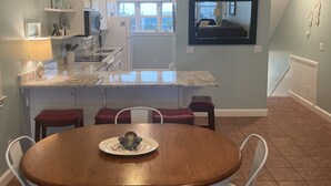 Una cuina moderna amb armaris blancs, una illa gran i una zona de menjador amb taula de fusta i cadires blanques.