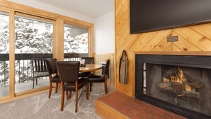 A cozy room with a fireplace, a dining table with chairs, and a large window overlooking a snowy landscape. - Cozy Slope-Side 2 Bedroom, Walk to Chairlifts! (Big Sky)