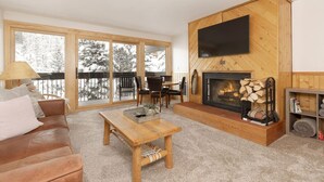 A cozy living room with a fireplace, a television mounted on the wall, and a balcony with a view of snow-covered trees. - Cozy Slope-Side 2 Bedroom, Walk to Chairlifts! (Big Sky)