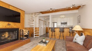 A cozy living room with a fireplace, a wooden coffee table, a brown sofa, and a TV mounted on the wall. - Cozy Slope-Side 2 Bedroom, Walk to Chairlifts! (Big Sky)