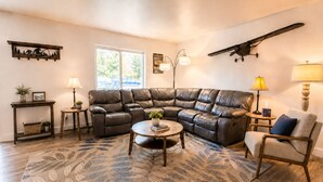 Uma sala de estar com sofá seccional, mesa de centro redonda, mesinha lateral, abajur, cadeira, objeto decorativo na parede e uma janela.