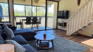 A spacious living room with a large sofa, a coffee table, and a TV mounted on the wall. There is a balcony with a view of trees and a ceiling fan.