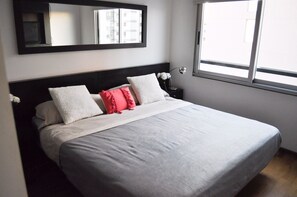 Un lit bien fait, avec une couverture grise et des oreillers, une table de chevet avec une lampe, et une fenêtre donnant sur des immeubles.