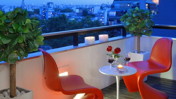 Un balcón con dos sillas rojas, una mesa pequeña con copas de vino y rosas, y vista a la ciudad al anochecer.