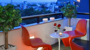 A balcony with two red chairs, a small table with wine glasses and roses, and a city view at dusk.