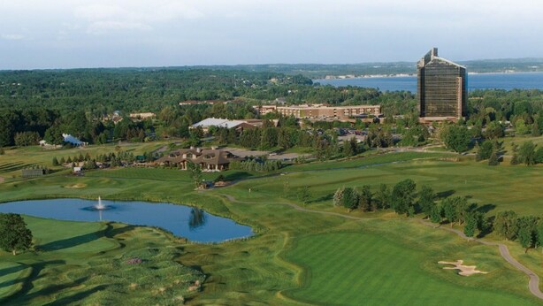 A golf course with a pond, a large building with a modern design, and a view of a body of water in the distance. - Spacious, minutes from Traverse City, Williamsburg a country setting. 2700 sq.  (Williamsburg)