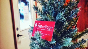 A small Christmas tree with a red sign labeled "The Little Village" in a room with a wooden door and a framed picture on the wall.