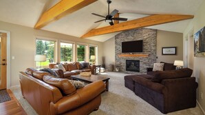 A spacious living room with leather sofas, a stone fireplace, and large windows. - 18 White Elm, 3 King Suites+Bunk Room! On 14th Green, Hot Tub, Bikes, 10 Passes (Sunriver)