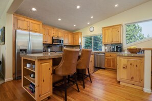 A kitchen with wooden cabinets, stainless steel appliances, and a breakfast bar with chairs. - 18 White Elm, 3 King Suites+Bunk Room! On 14th Green, Hot Tub, Bikes, 10 Passes (Sunriver)