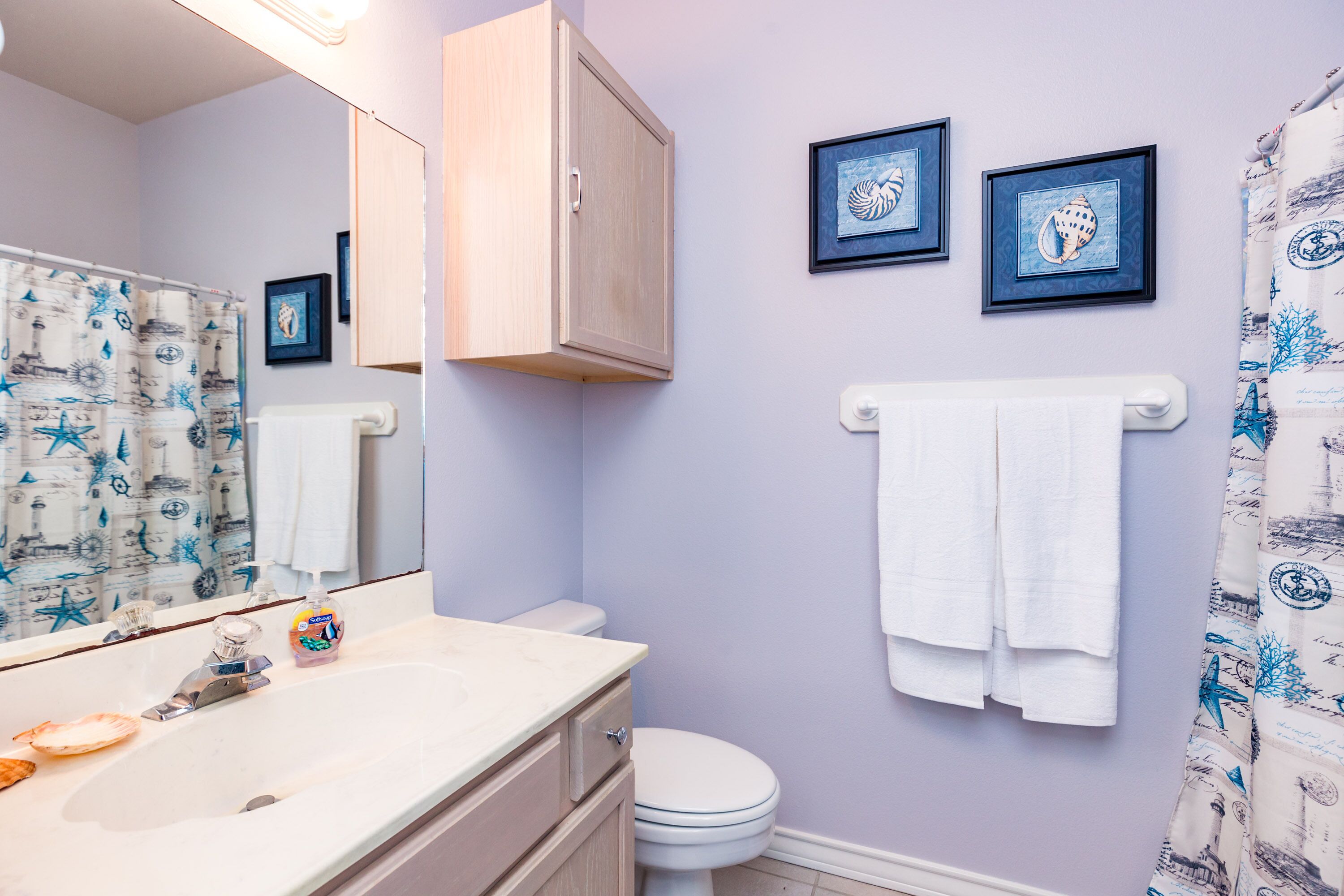 Ein Badezimmer mit einem weißen Waschbecken, einer Toilette und einem Duschvorhang mit maritimen Motiven. An der Wand hängen zwei Bilder und ein Handtuchhalter mit zwei weißen Handtüchern.