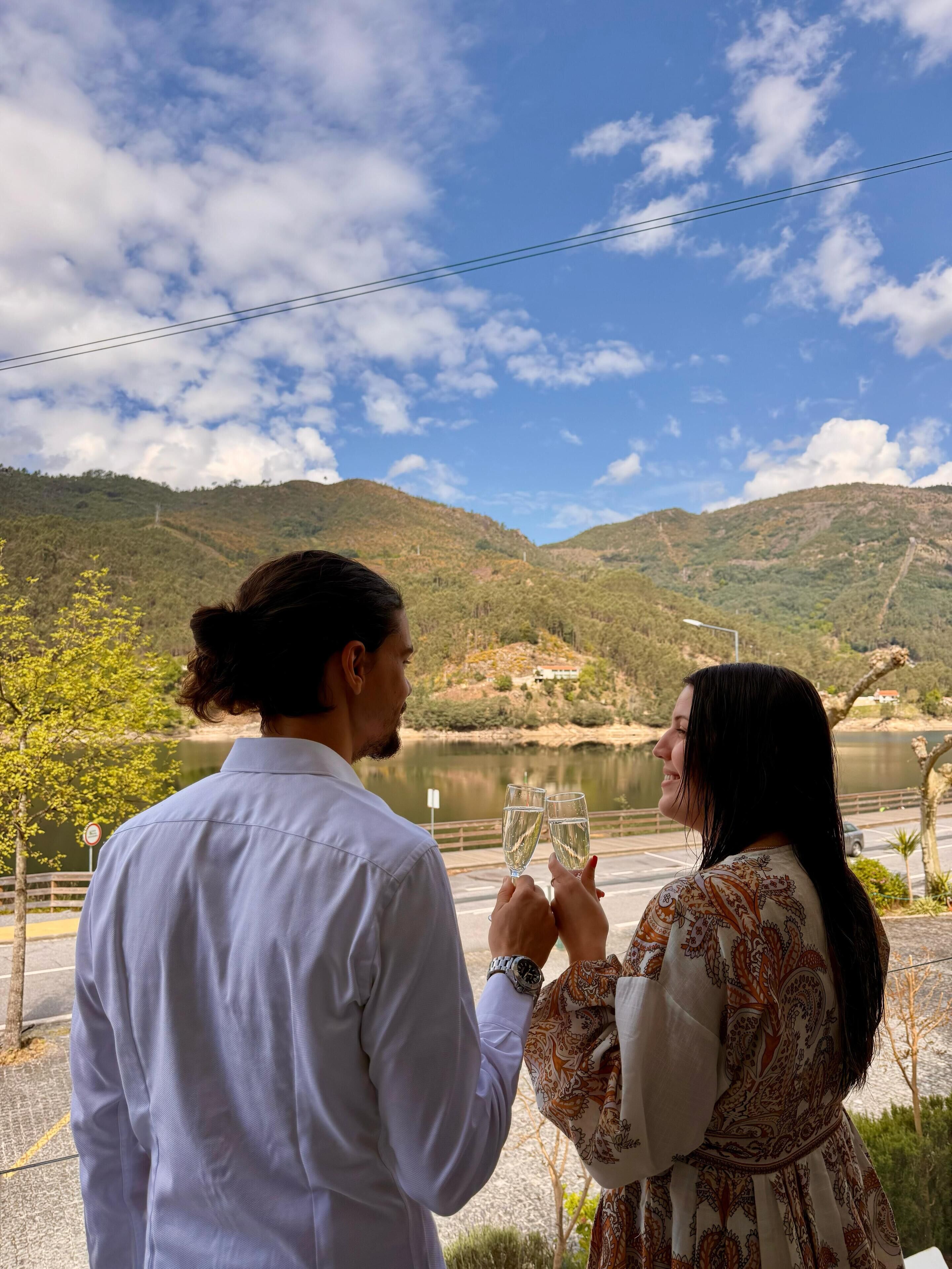 Deux personnes tenant des verres, avec une vue sur un lac et des collines en arrière-plan.