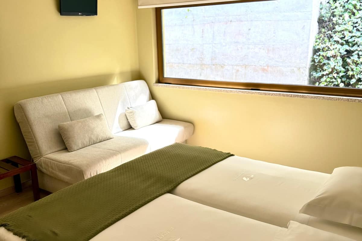 A hotel room with two beds, a sofa, a TV, and a window with a view of greenery.