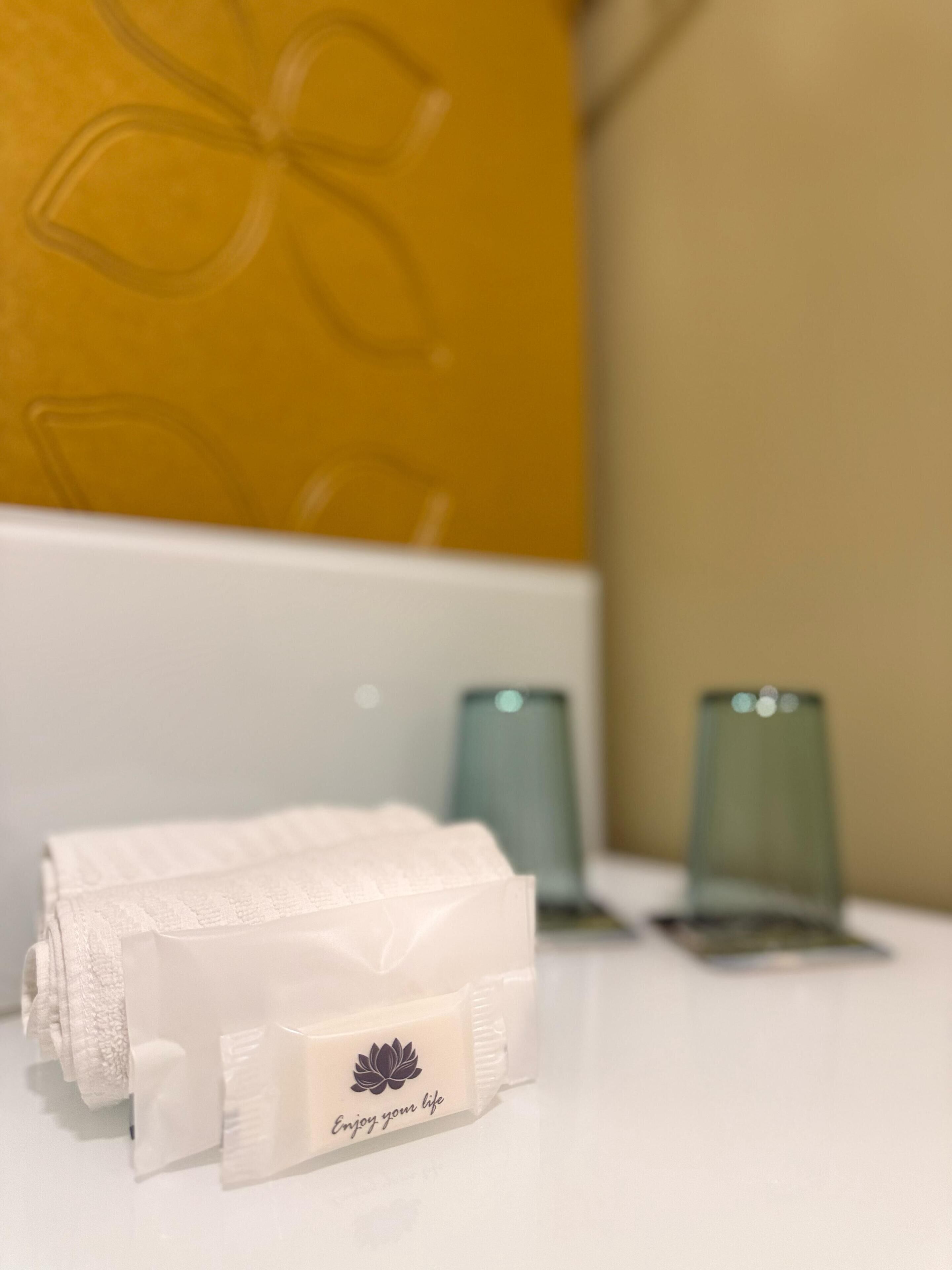 A neatly folded white towel with a lotus flower logo and the text "Enjoy your life" on a white surface, with a yellow pillow and two green glasses in the background.