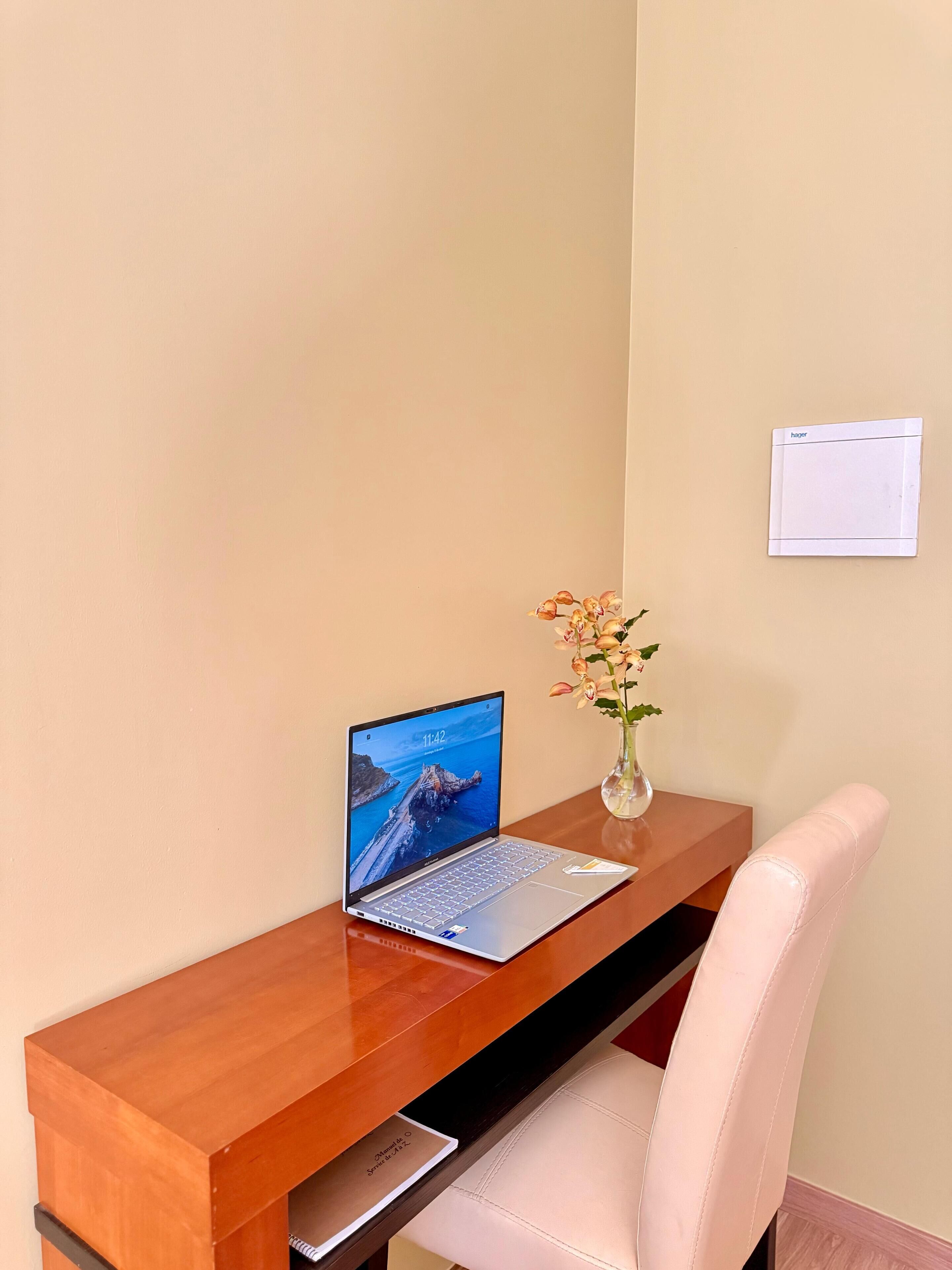 A wooden desk with a laptop displaying a scenic wallpaper, a vase with flowers, and a white chair.